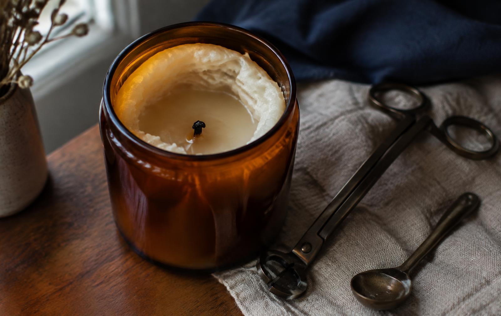 A close-up of an amber glass candle with wax tunneling down the middle, beside a wick trimmer and a wick dipper on a linen cloth